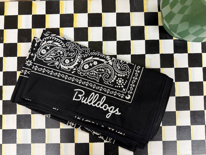 A black bandana-style cloth with white paisley print and the word 'Bullocks' in script, laid flat on a black-and-white checkered surface.