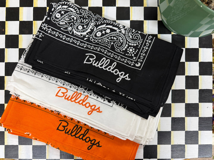Three folded bandanas in orange, white, and black are arranged on a black and white checkered surface. The black bandana features a white paisley border and the word 'Bulldogs' in white script, while the other two show different colored script versions of the same word.