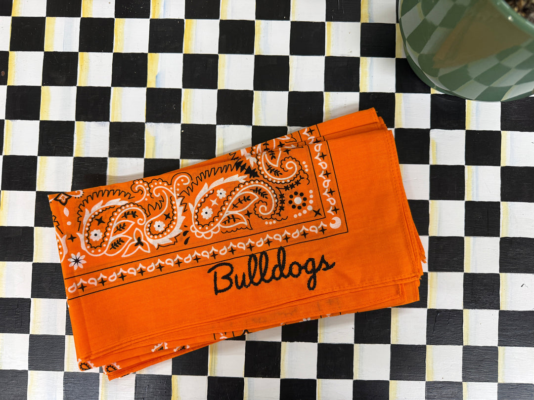 An orange bandana-style cloth napkin with a white paisley print and the word 'BULLDOGS' in black script, laid on a black-and-white checkered surface.