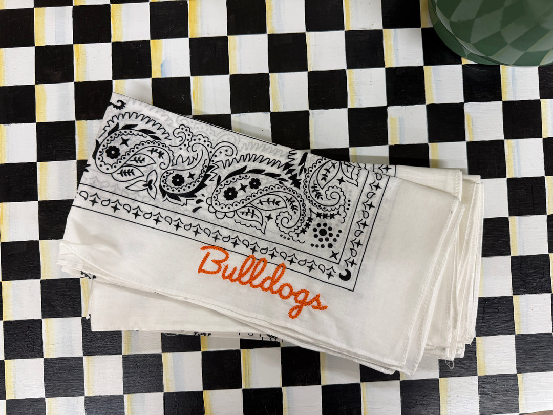 A stack of white cotton bandanas featuring a black paisley border design, one with orange script text reading 'Bulldogs', and another with 'Clemson' in a decorative font, laid flat on a black and white checkered surface.