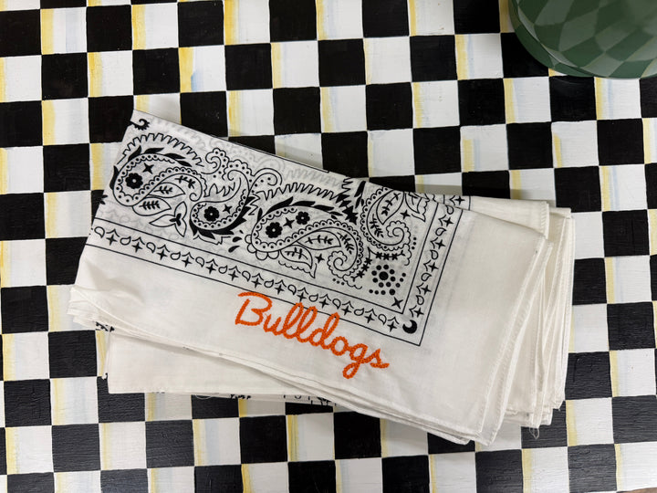 A stack of white cotton bandanas featuring a black paisley border design, one with orange script text reading 'Bulldogs', and another with 'Clemson' in a decorative font, laid flat on a black and white checkered surface.